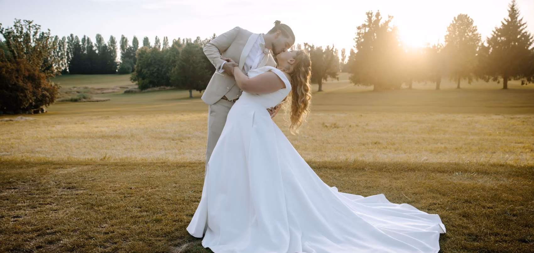 Couple de mariés s'embrassant dans un parc au coucher du soleil, la traîne de la robe déployée dans l'herbe