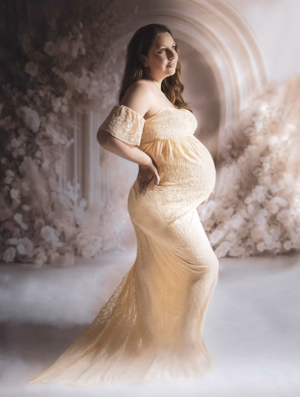 Future maman en robe de dentelle crème, posant de profil devant un décor floral en studio