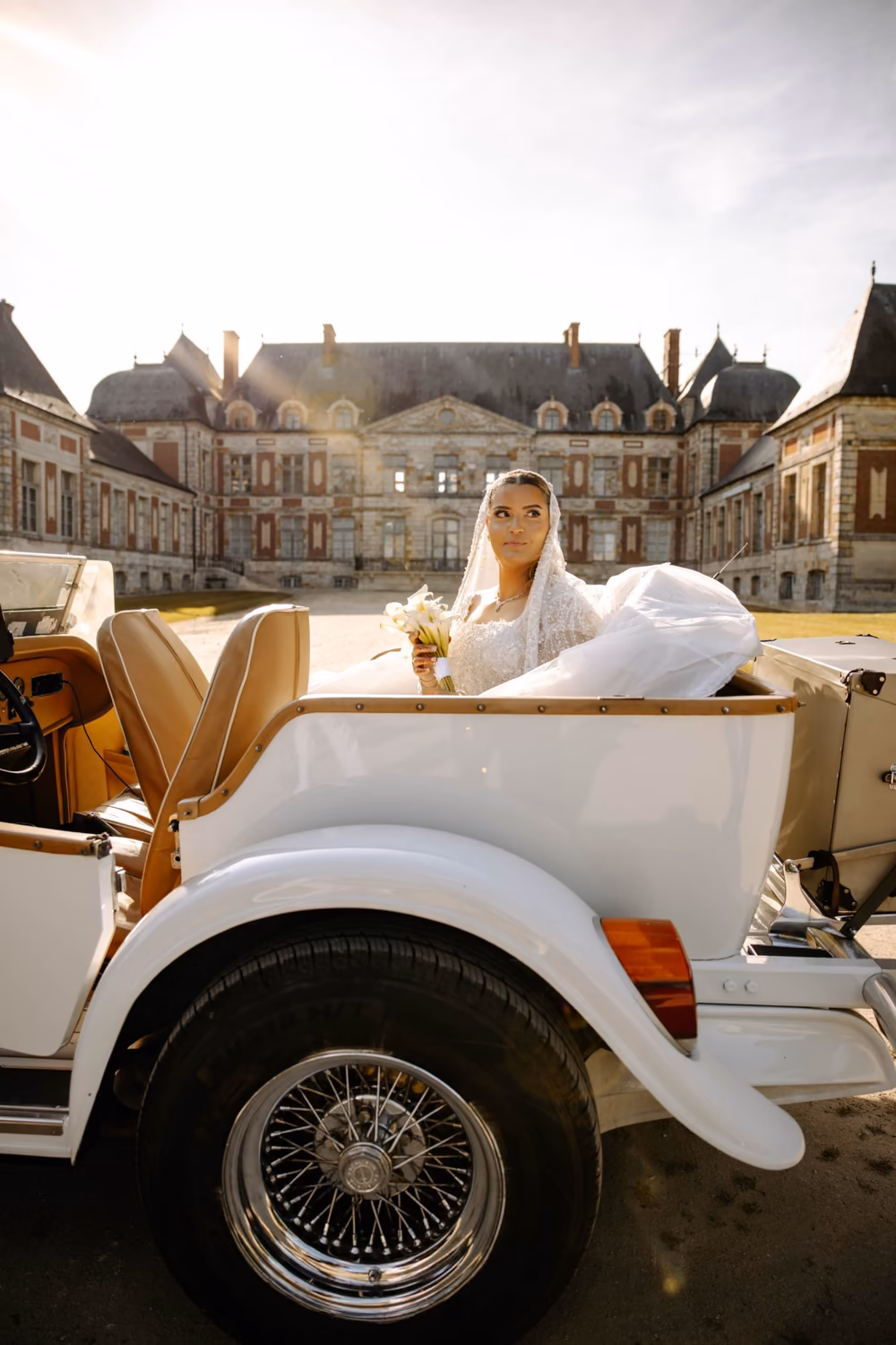 Mariée en robe brodée et voile de perles, assise dans une voiture de collection blanche devant un château
