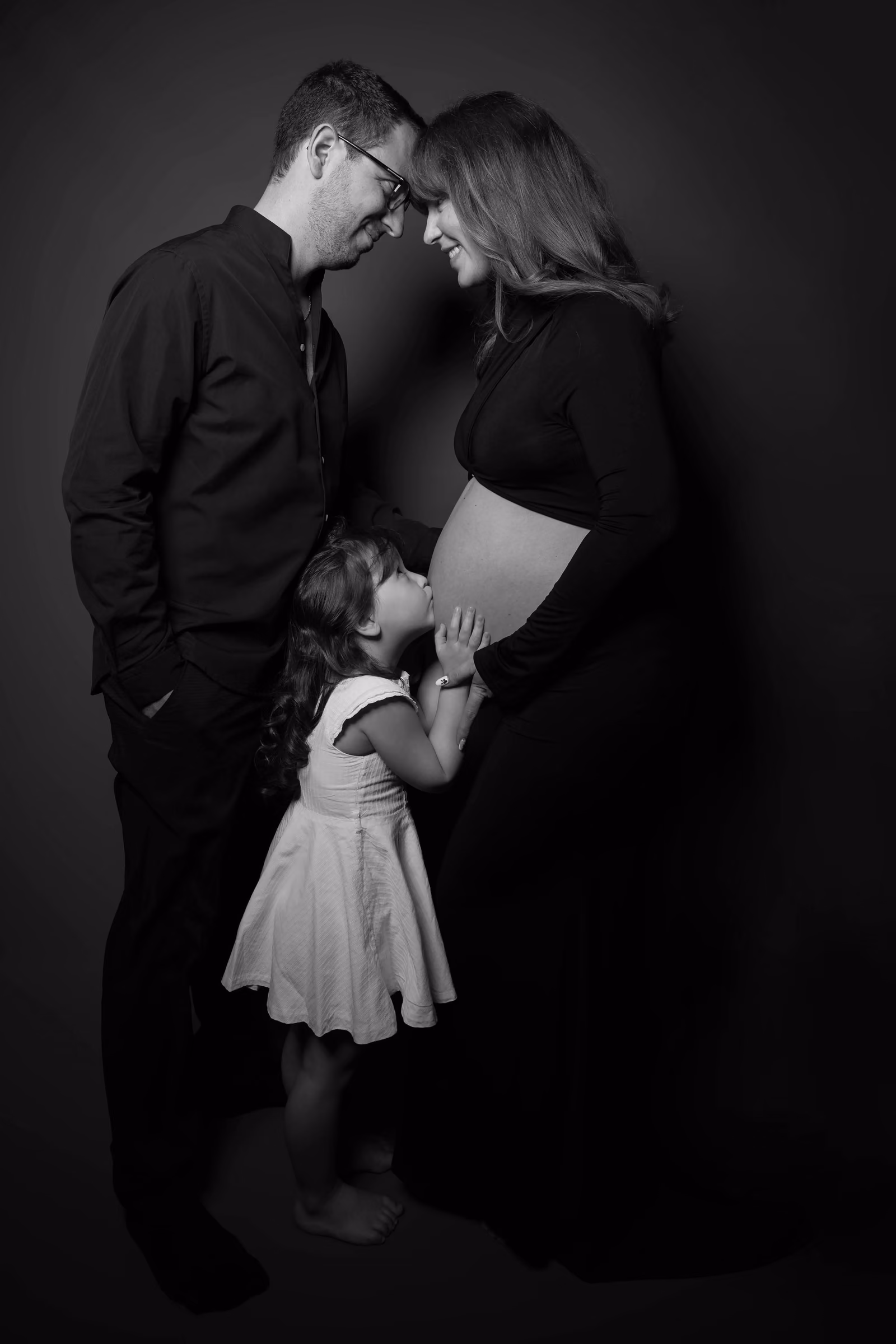 En noir et blanc, une petite fille embrasse le ventre de sa maman enceinte, le papa penché au-dessus