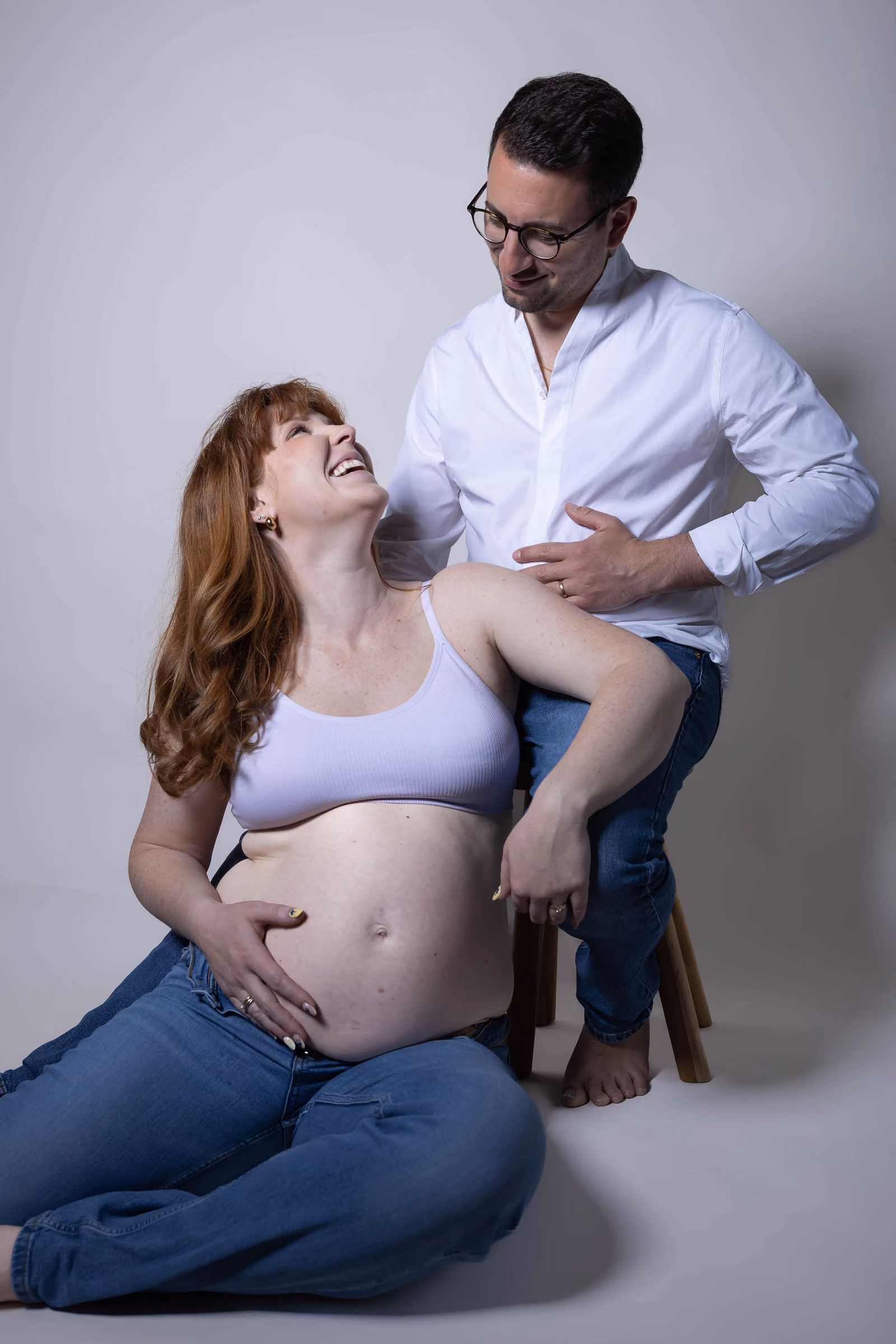La future maman assise au sol éclate de rire, le papa penché derrière elle