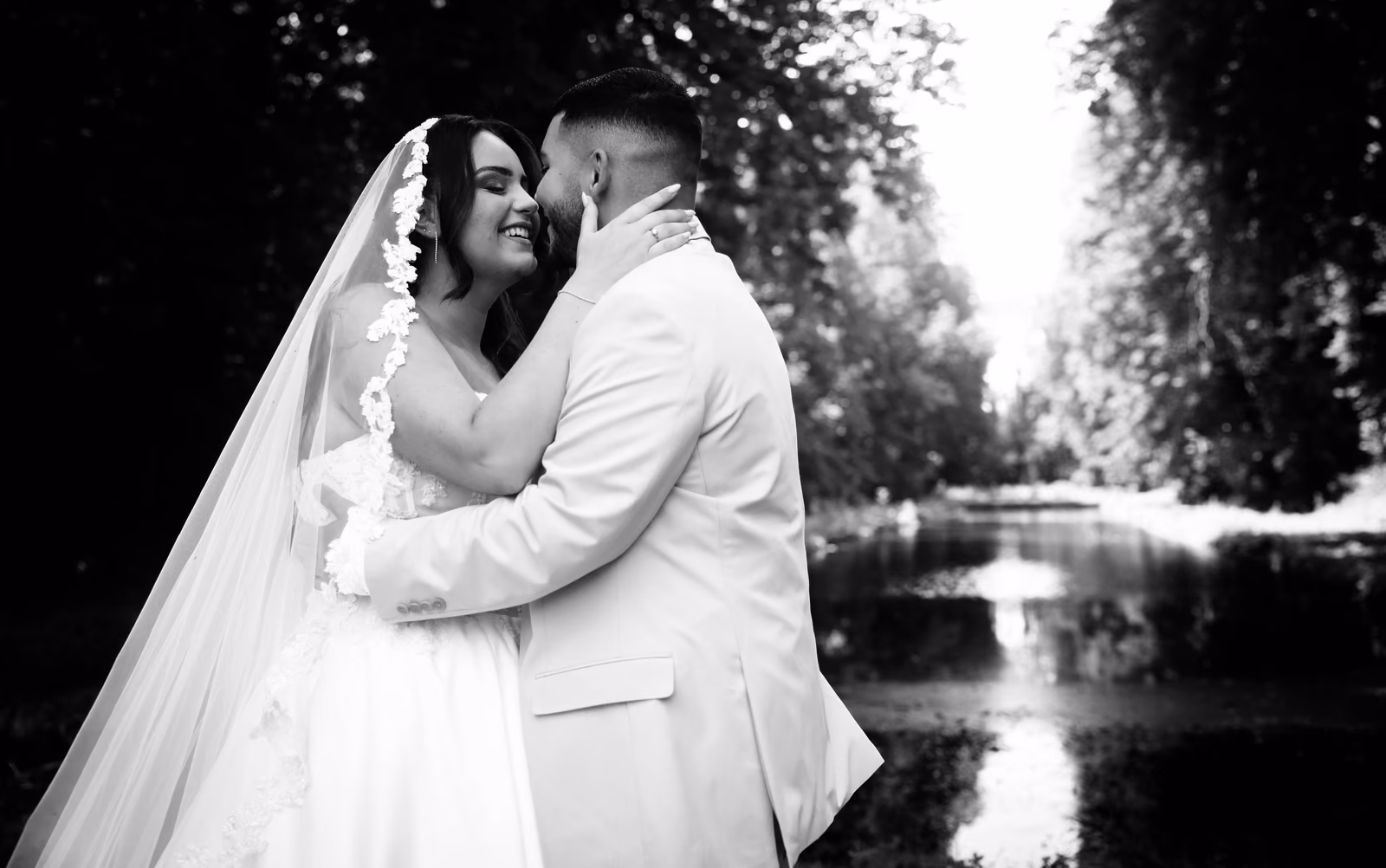 En noir et blanc, les mariés joue contre joue au bord d'un miroir d'eau bordé d'arbres