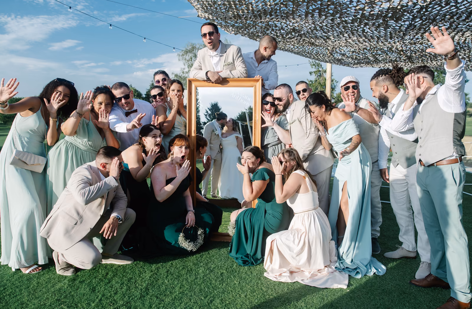 Photo de groupe joyeuse sous une voile d'ombrage, les invités en vert d'eau autour d'un cadre doré