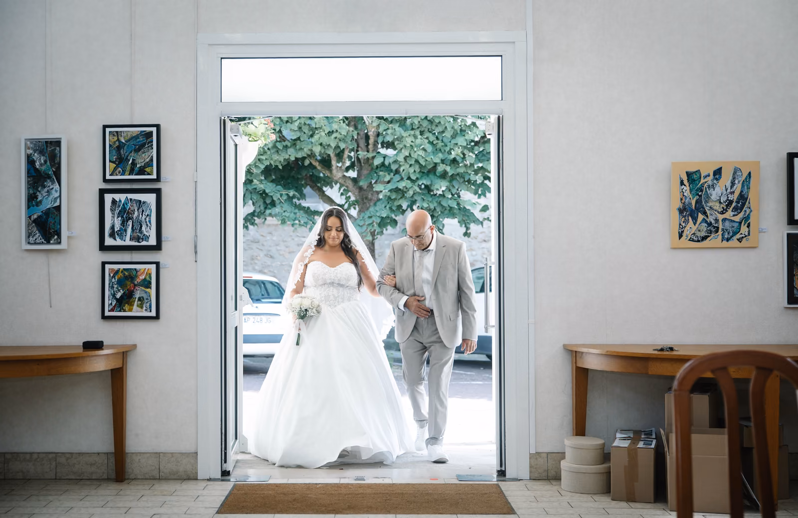 La mariée au bras de son père, en contre-jour dans l'embrasure d'une porte vitrée