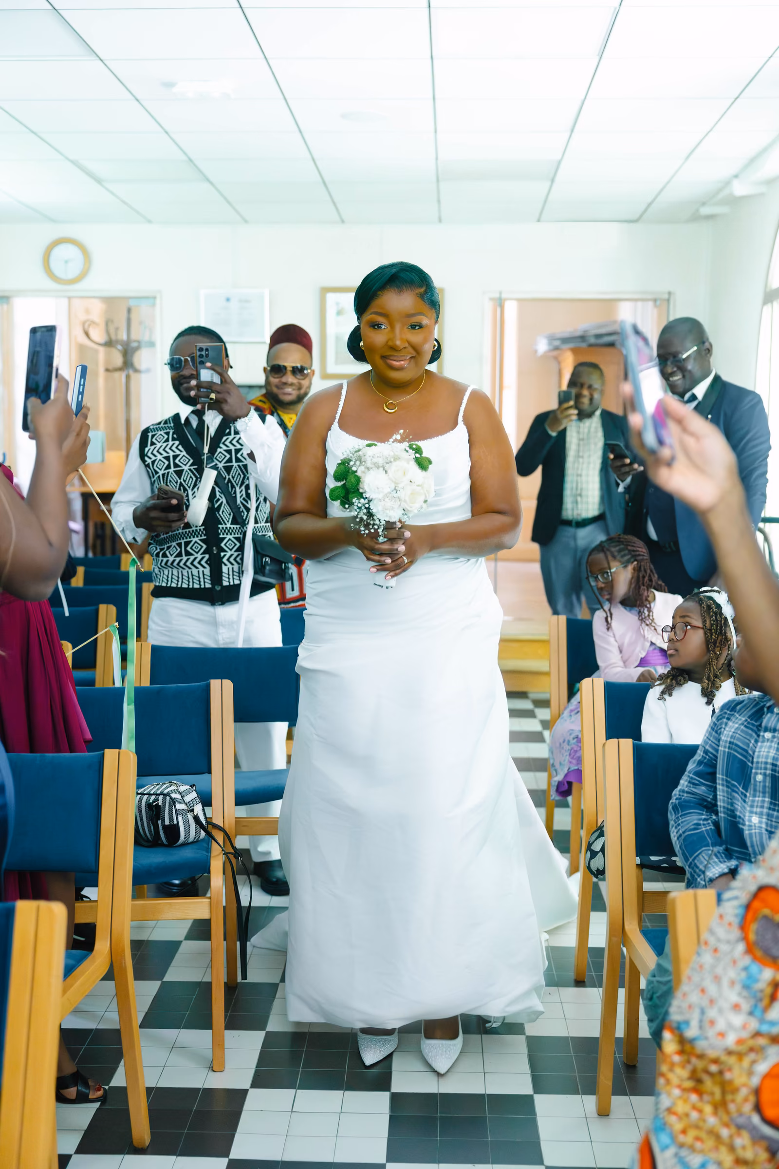 La mariée remonte l'allée de la salle de cérémonie, bouquet à la main, sous les téléphones des invités
