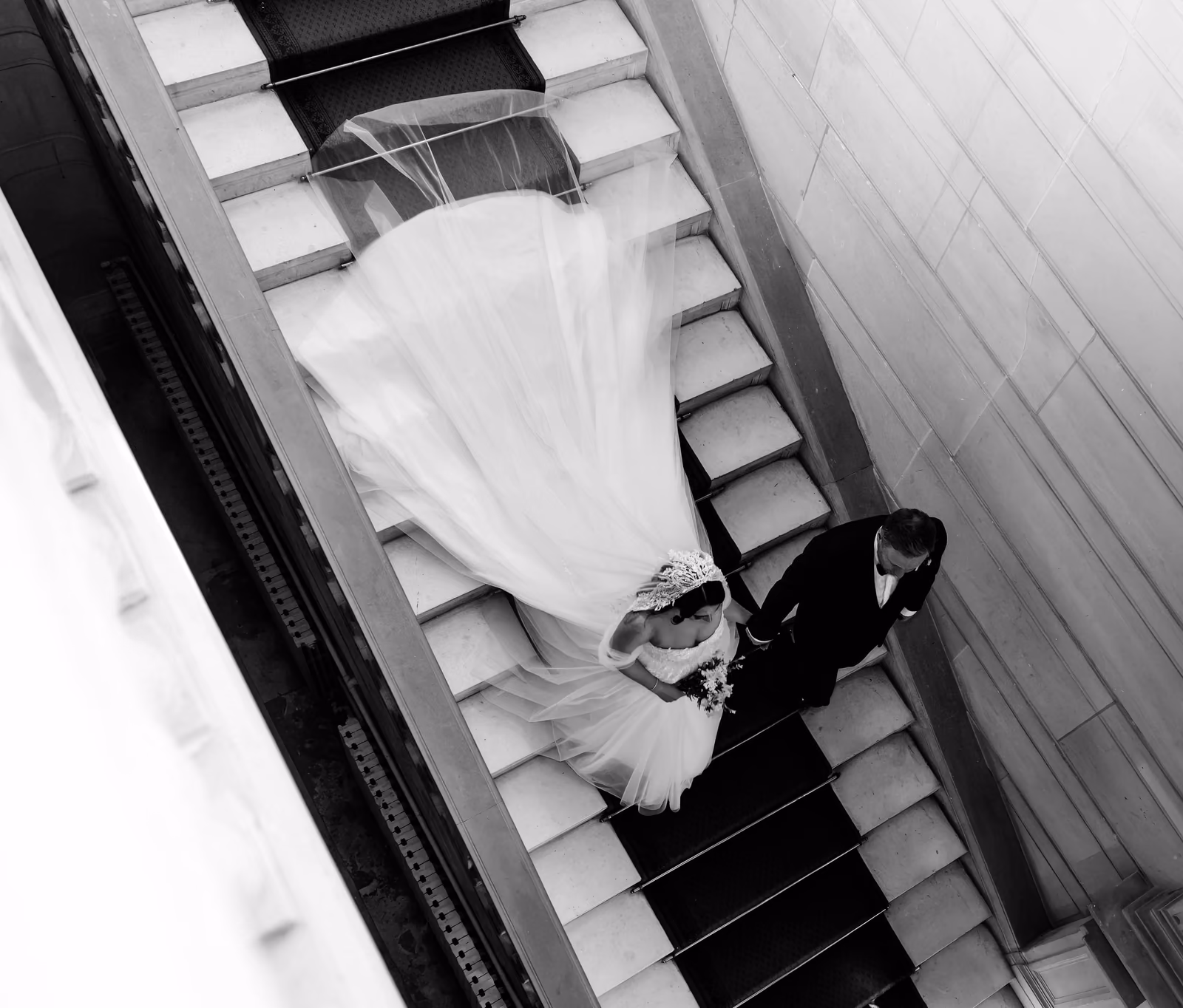 Vue plongeante en noir et blanc sur les mariés dans l'escalier, la traîne en éventail