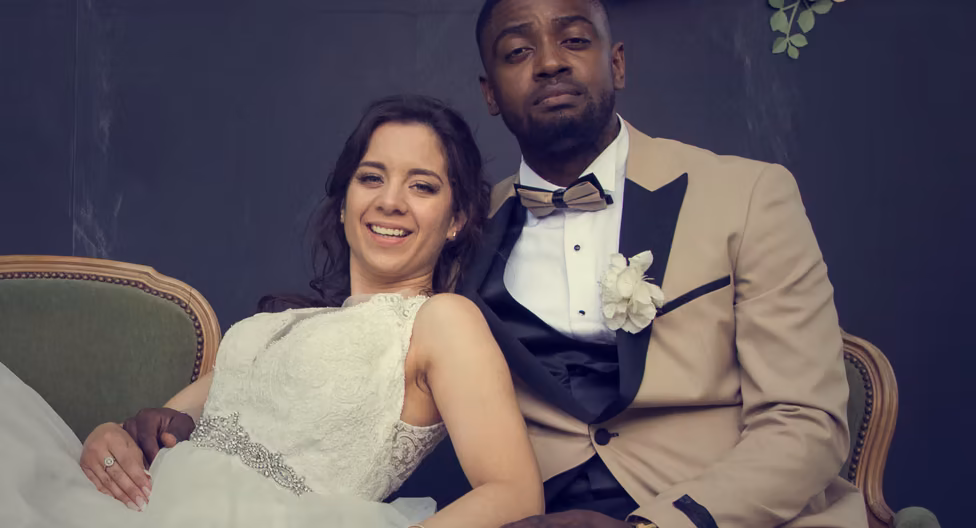Couple de mariés assis côte à côte sur un canapé de velours vert, elle en robe brodée, lui en costume beige et nœud papillon